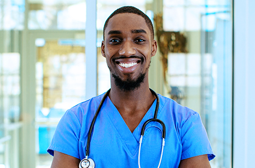 Male healthcare provider dressed in scrubs, facing the camera and smiling in front of a glass wall.