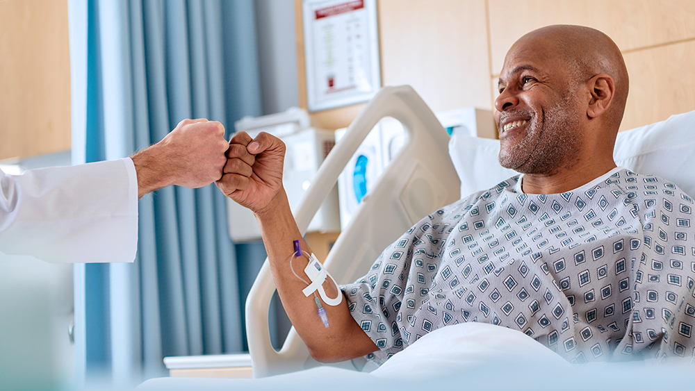Patient in a hospital bed has the Clik-FIX securement device in his arm as he fistbumps a provider.