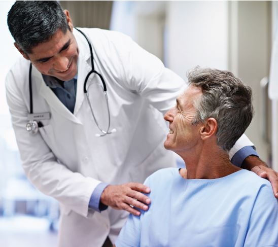 Doctor leaning over a patient with one arm placed on the patient's shoulder as they both smile at each other. 
