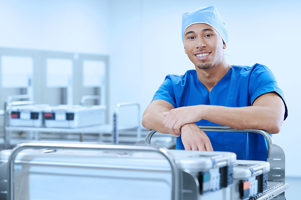 Man in scrubs smiling while standing in the OR and leaning on a cart with containers placed on it.