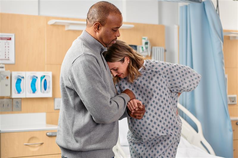 A person in a hospital gown being supported by another person next to a hospital bed with medical equipment in the background.