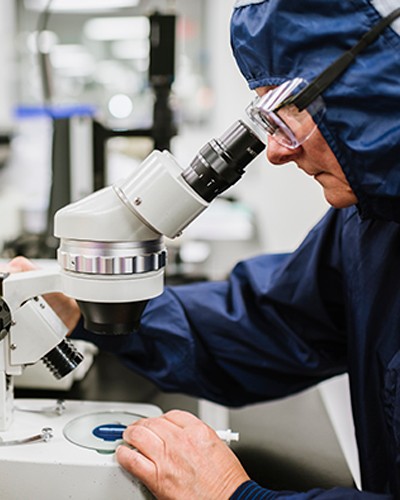 Employee with safety gear on looking into a microscope analyzing data. 