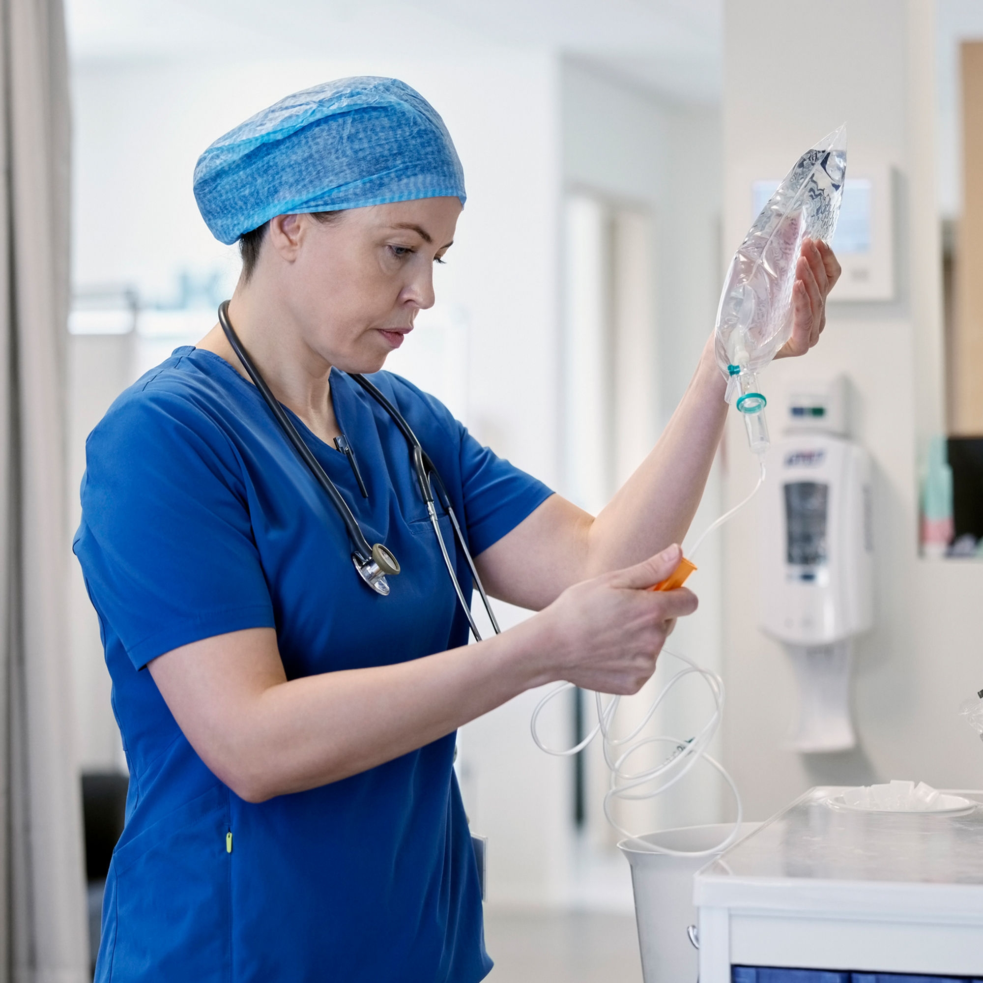 A female nurse inspects an IV bag set in a non-descript hospital room.