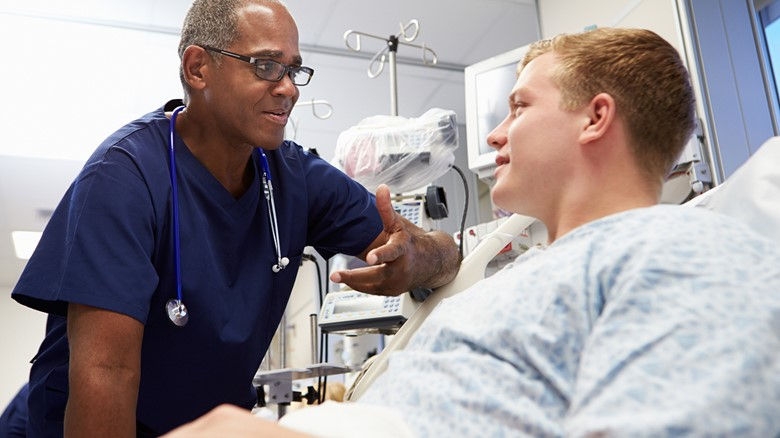A middle-aged male healthcare professional talks to a younger male patient in a hospital bed with medical equipment in the background.