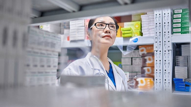 A female pharmacist inspects shelves lined with various kinds of medications in a hospital pharmacy setting.