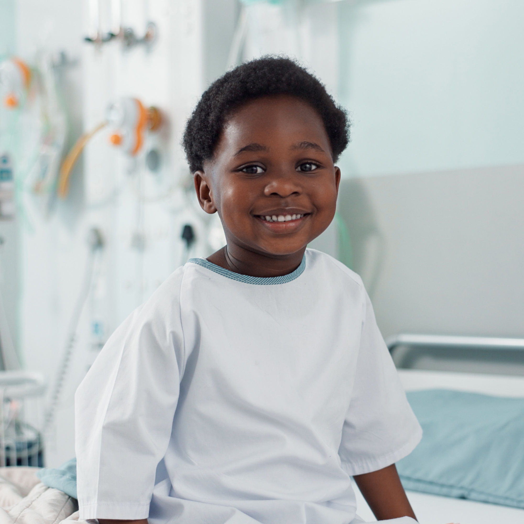 A little boy in hospital garb sits on a hospital bed with an array of medical equipment—including pumps, wires and tubes.