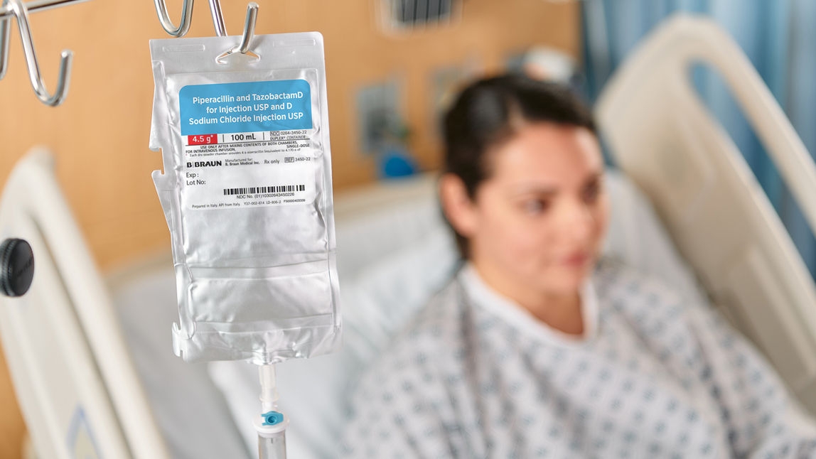 A 100mL bag of B. Braun’s Piperacillin and Tazobactam hangs from an IV pole in the foreground while a female patient reclines in a hospital bed in the background.