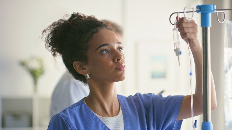 A female nurse inspects IV equipment hanging from an IV pole in a care setting. 