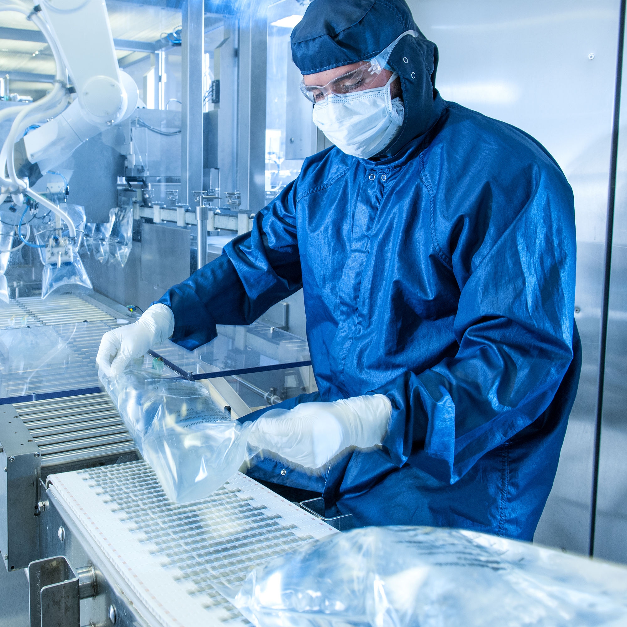 A worker wearing head-to-toe protective gear inspects IV bags on a conveyor belt in a sterile manufacturing facility. 