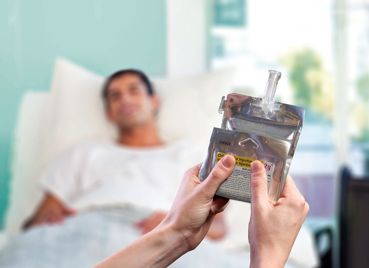 In the foreground, two hands hold a folded flexible IV container while a patient reclines in a hospital bed, out of focus, in the background.