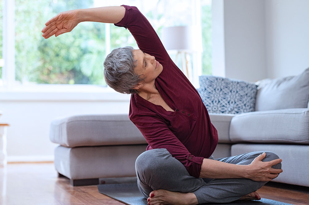 Woman sitting on the floor in front of a couch doing yoga, stretching with her arm over her head. 