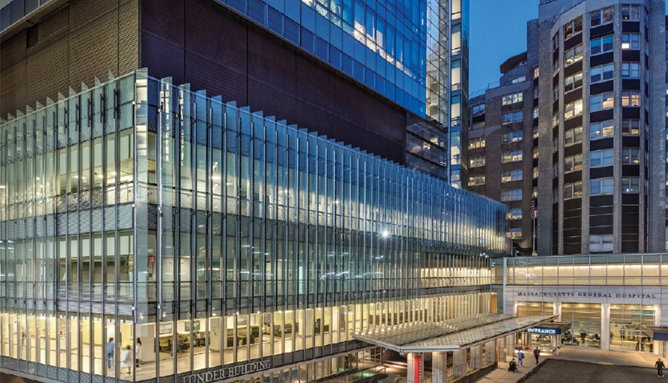 An exterior shot of the Mass General Hospital building in Boston during the evening. The building has large glass windows that make up the first few floors, a floor without windows and the rest of the floors seen are all windows lit up.