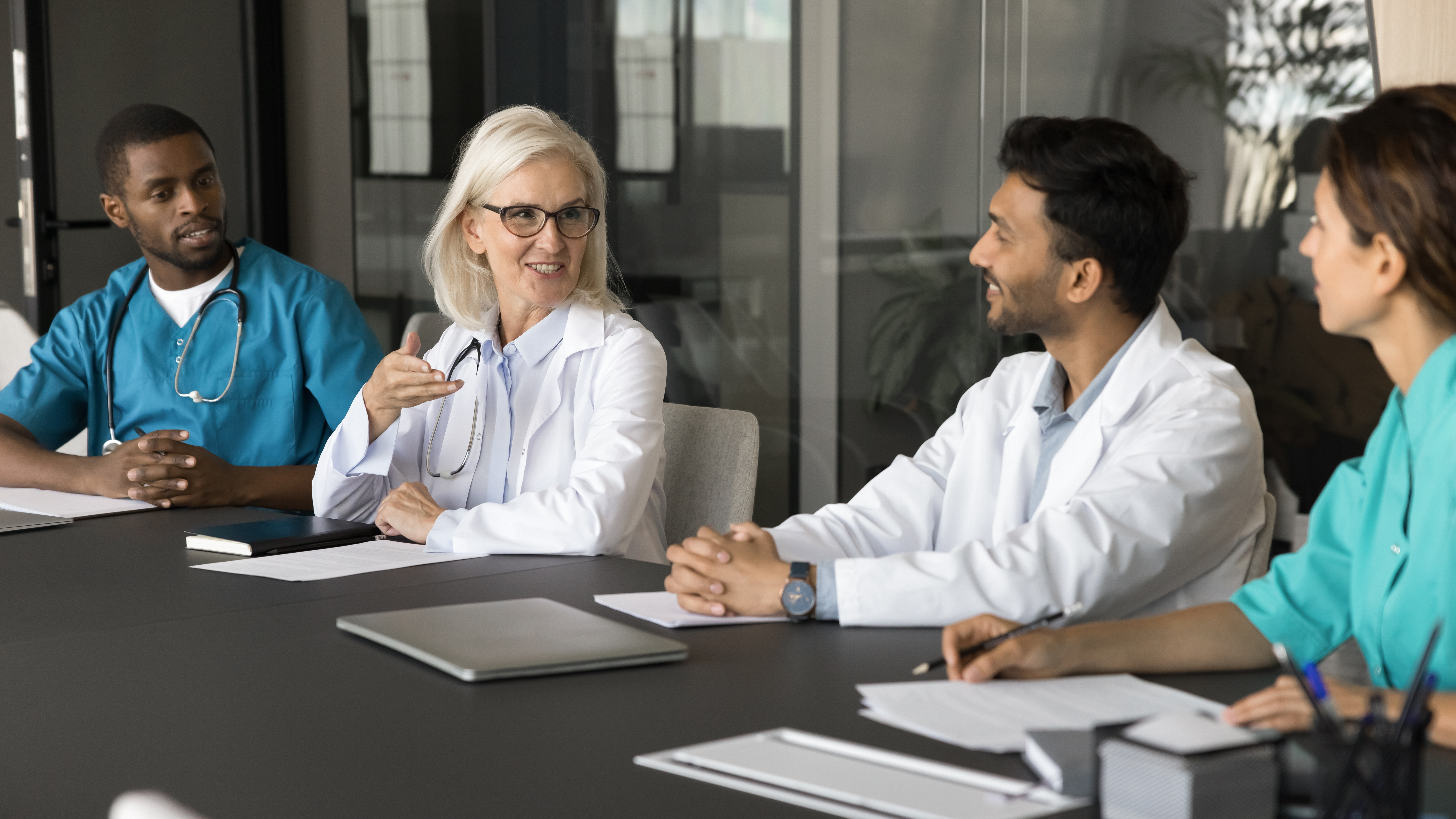 Healthcare professionals seated at a table in discussion, with a laptop and documents visible.