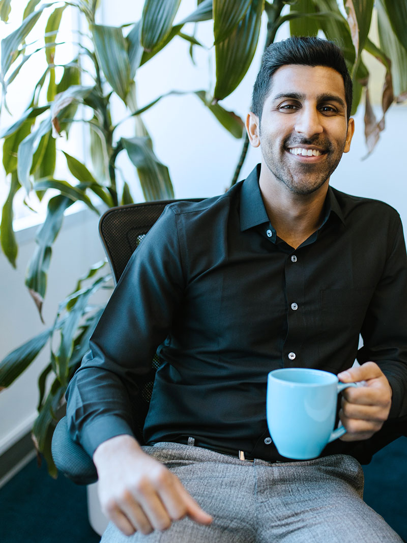 Professionally dressed man holding a mug, smiling at the camera, while sitting in an office chair with a plant in the background. 