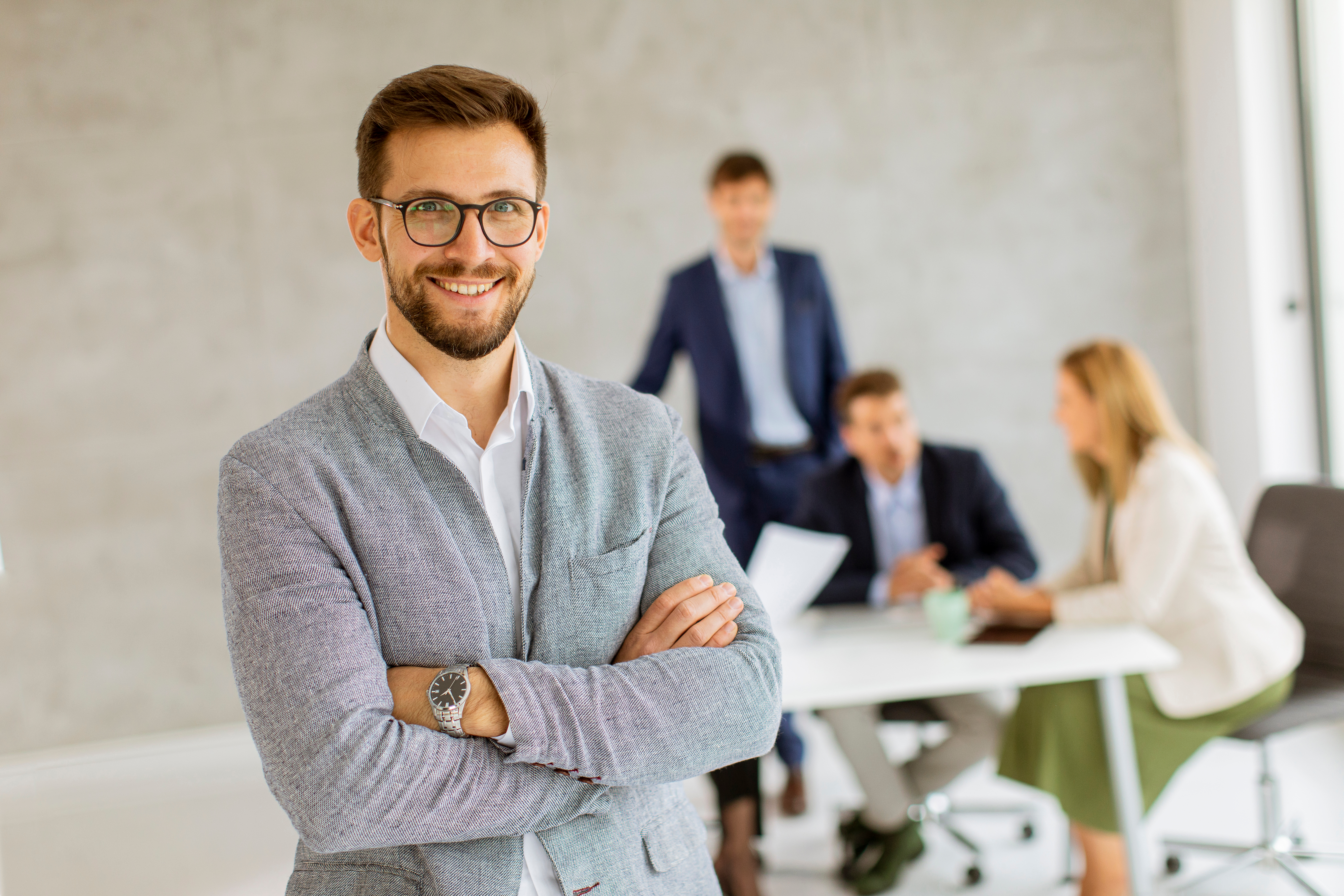 Businessman with arms crossed smiling in an office setting with colleagues blurred in the background behind him.
