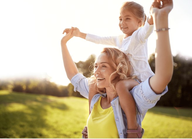 A woman has a young girl sitting on her shoulders as they smile while standing in a grassy field.