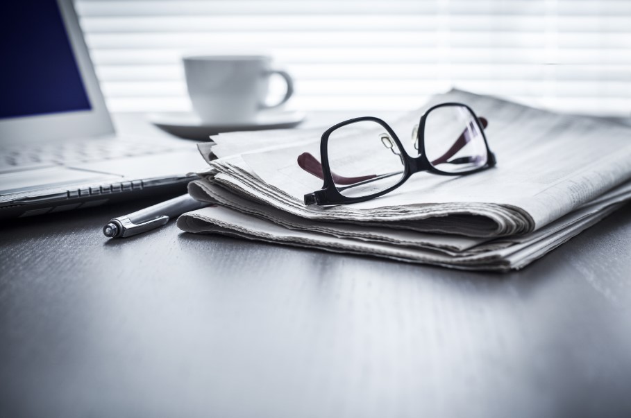 A table hold eye glasses resting on a newspaper with a laptop and coffee cup in the background.