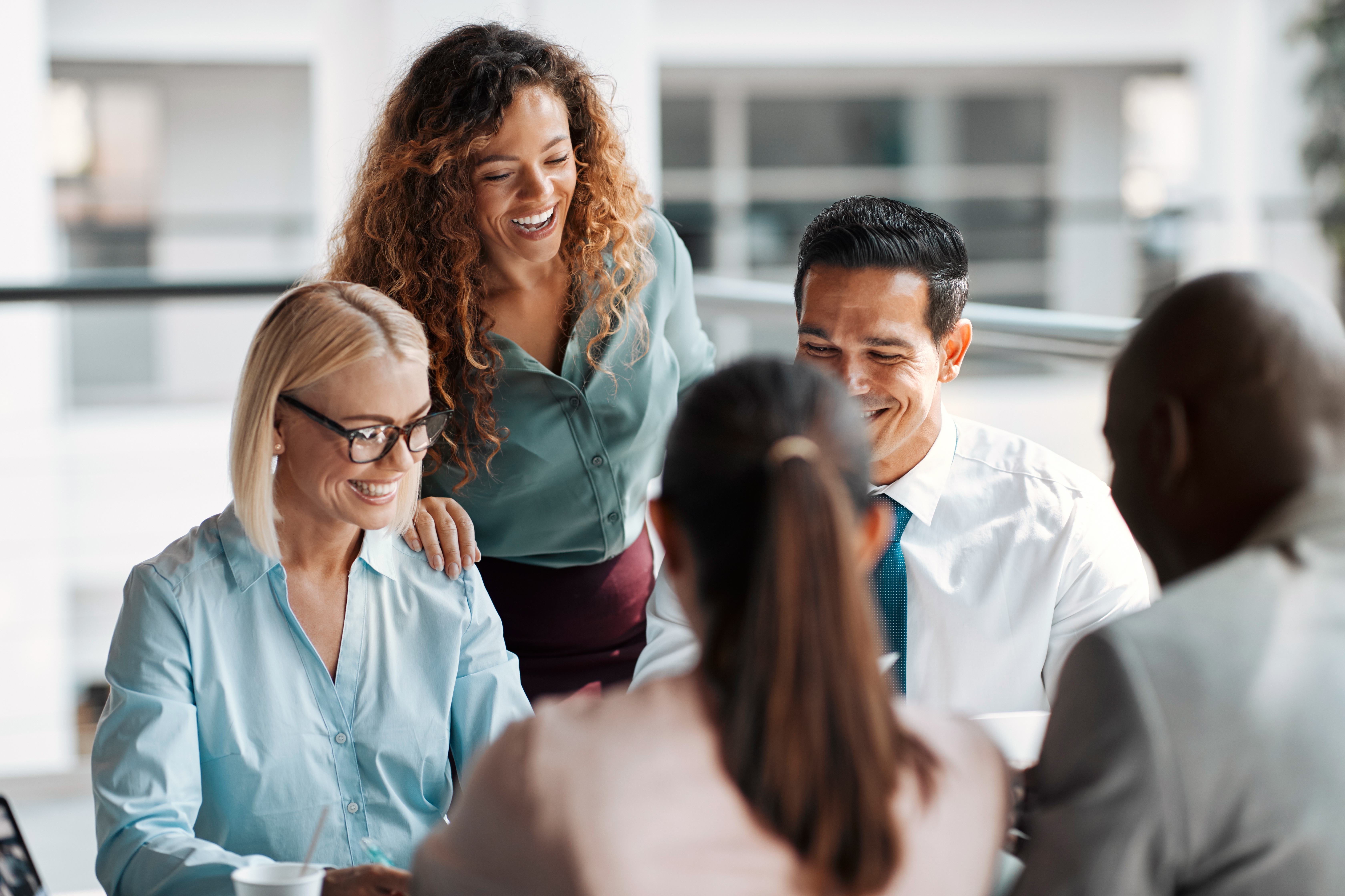 A group of individuals in a work setting, gathered around a table while talking and smiling.