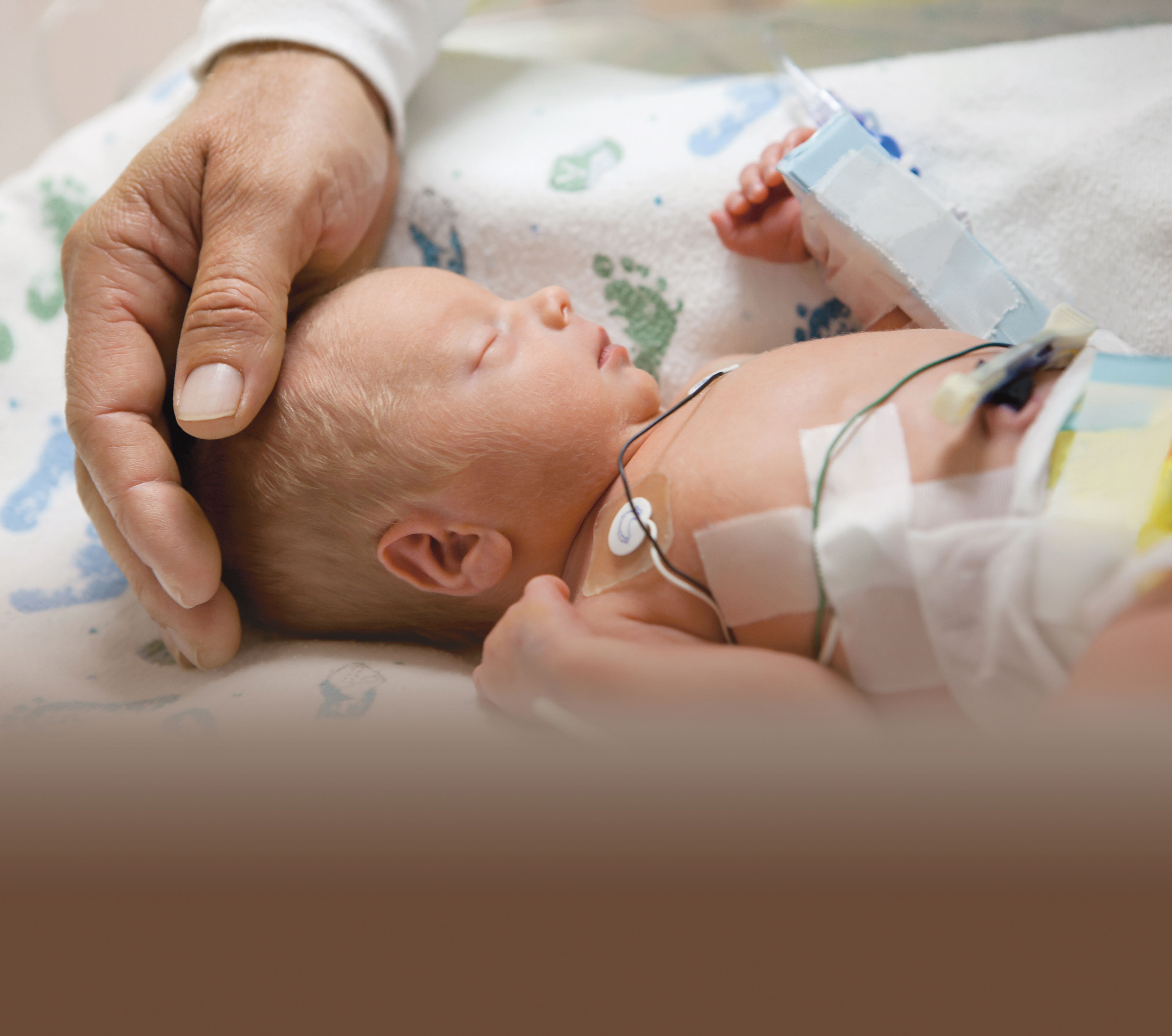 Small baby sleeping while in a hospital setting with a gentle adult hand resting on the baby's head.