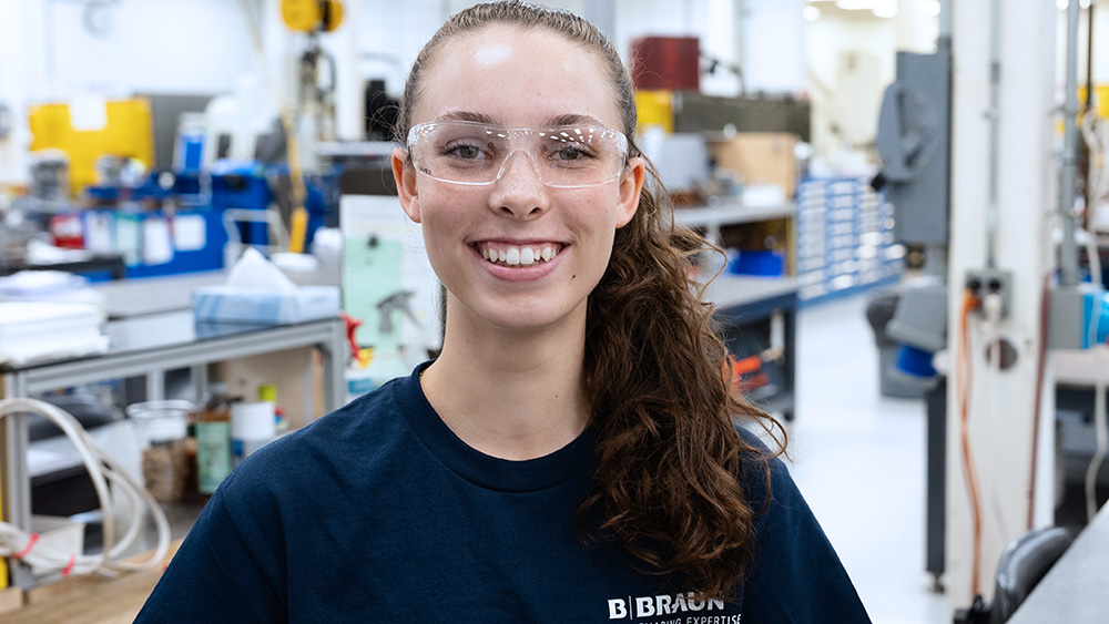 B. Braun employee wearing safety glasses smiling toward a camera while standing in a manufacturing setting.