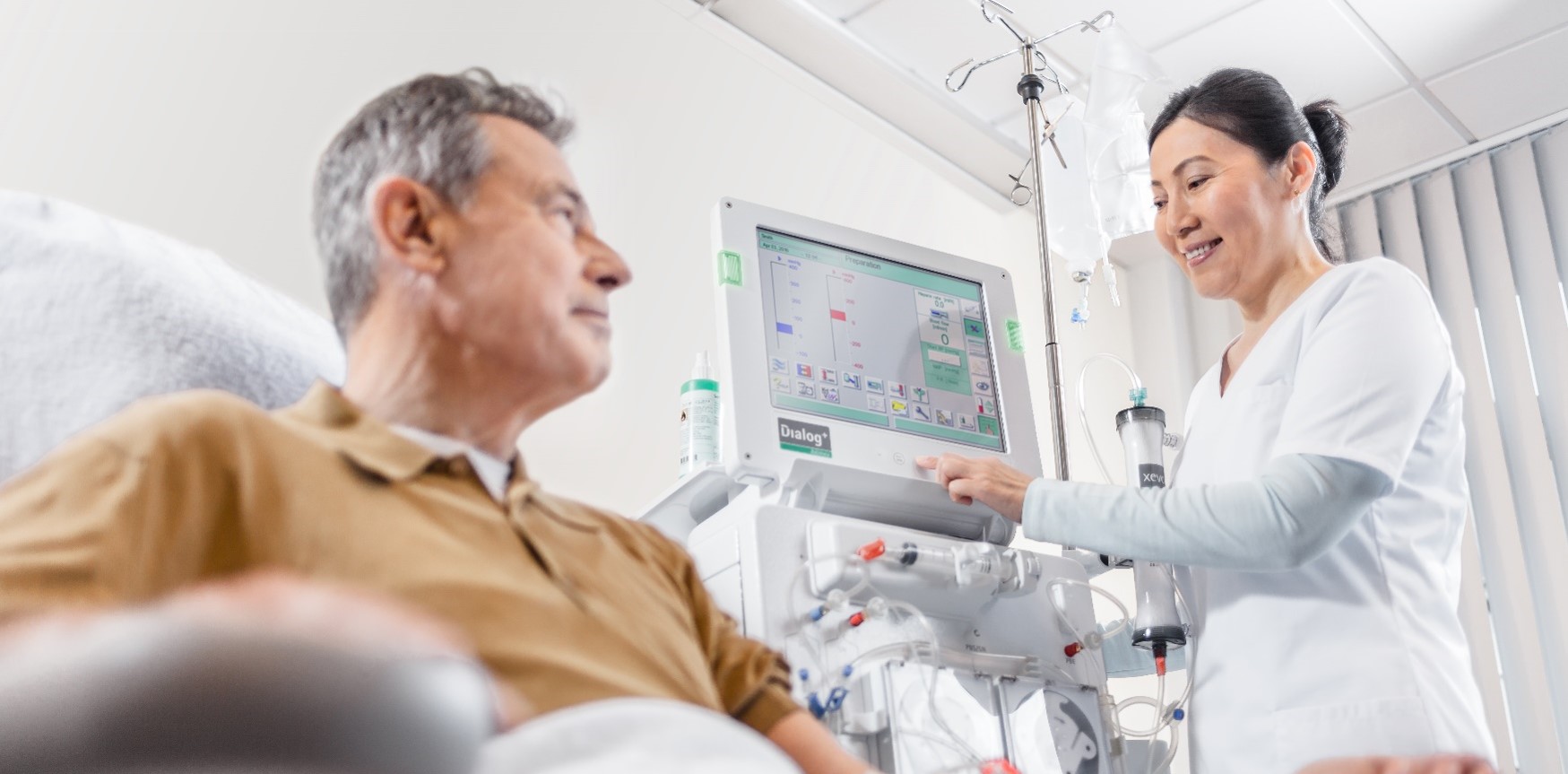 Patient sitting in a chair in a hospital setting as a nurse stands beside him, programming a B. Braun Dialog + Hemodialysis Machine to begin the dialysis treatment.