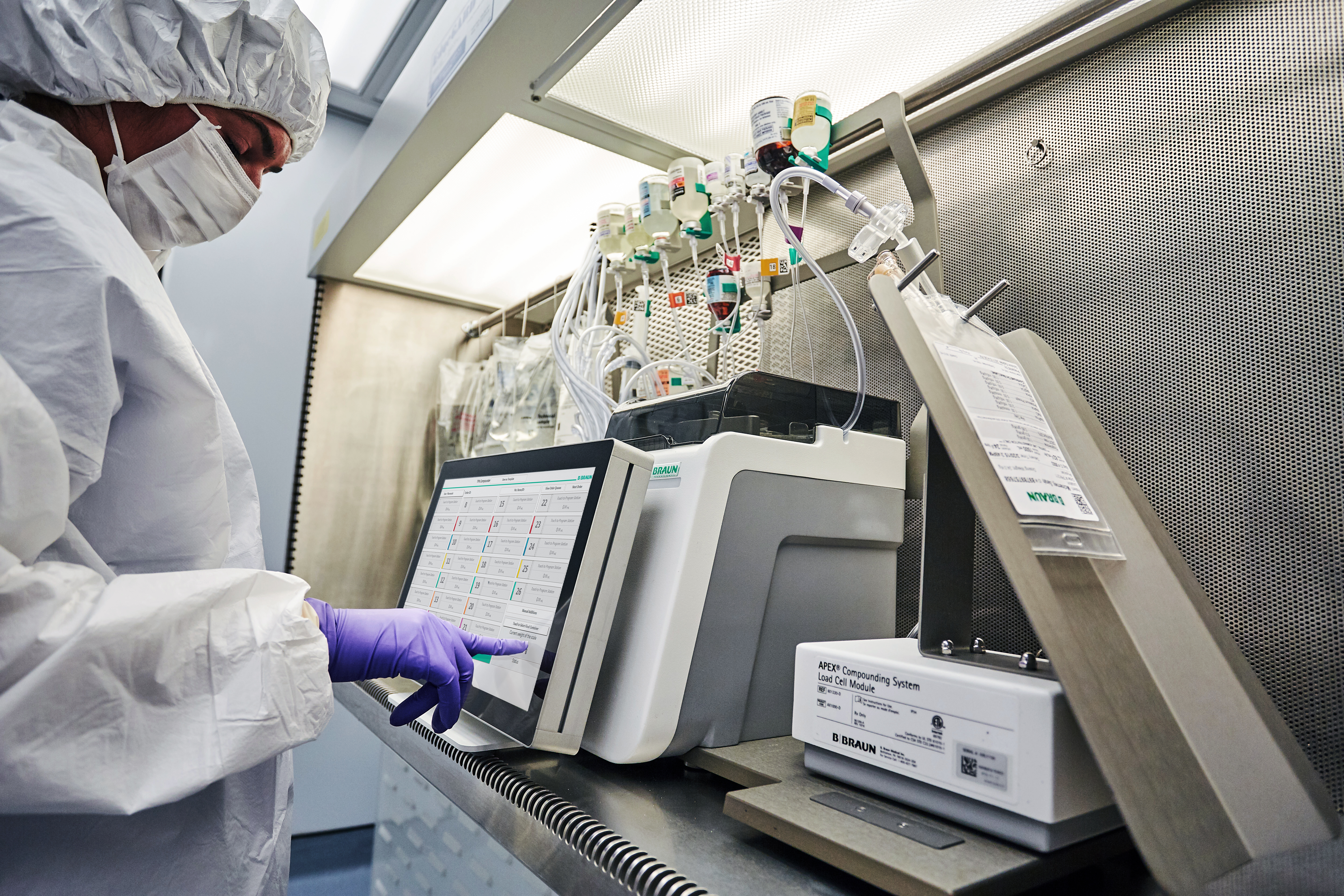 A person in protective clothing reaches out a purple gloved hand to touch the screen of the APEX Compounding System.