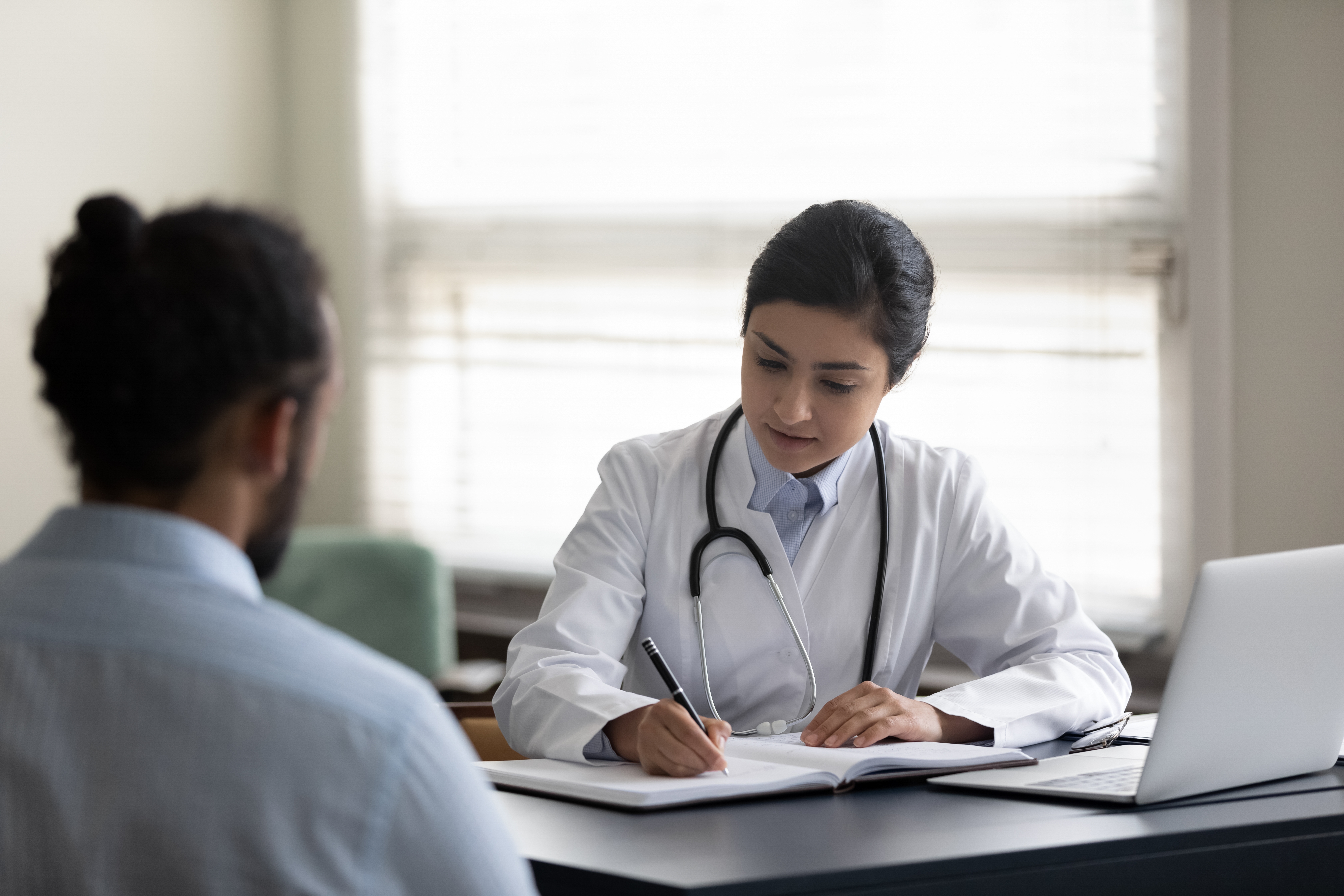 A healthcare provider in an office setting sits across from a patient and writes in a notebook. The patient has his back to the camera and is out of focus.