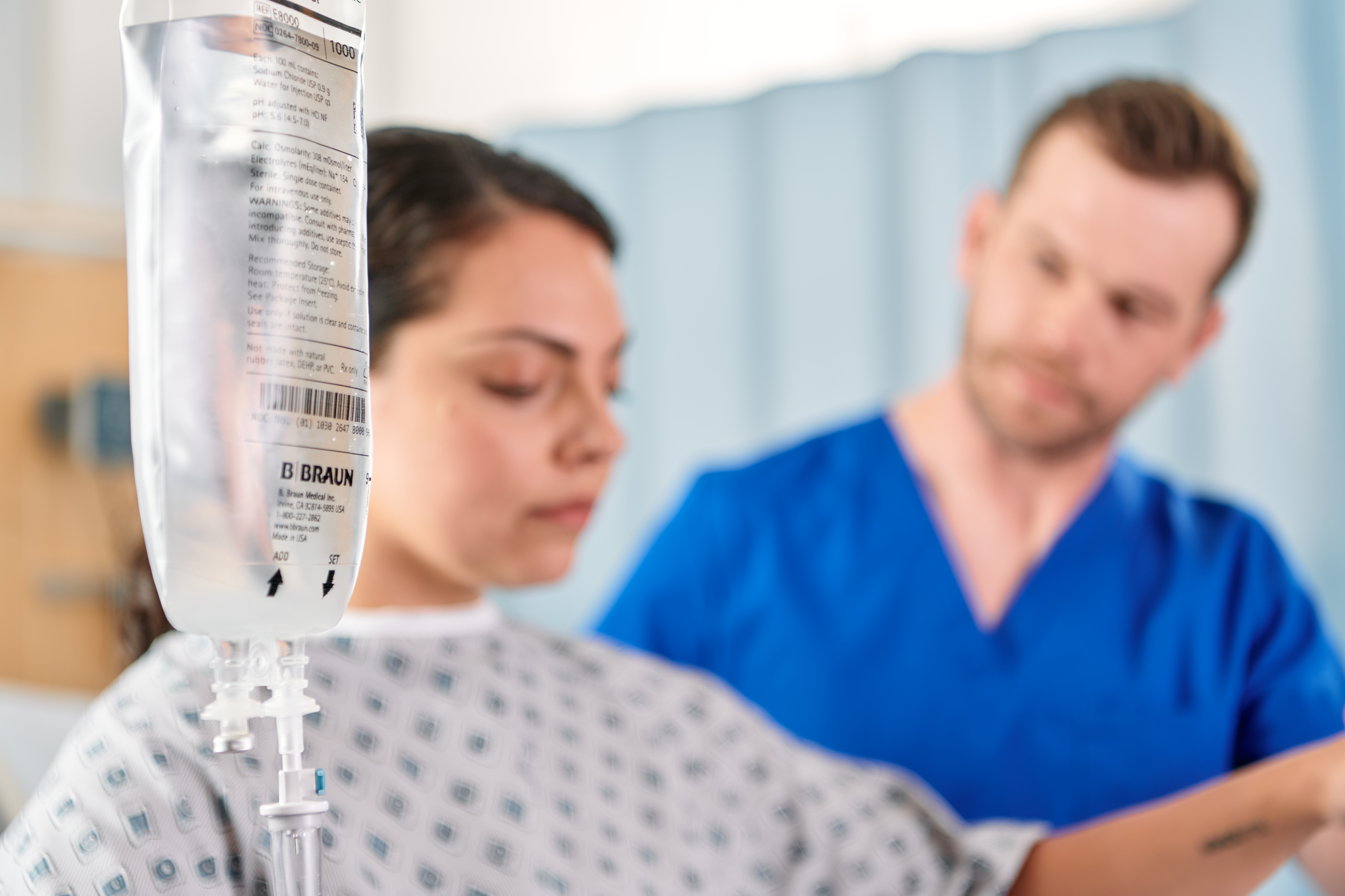 In the foreground, a B. Braun IV bag hangs, while out of focus in the background a patient stands with her eyes shut and one arm out while a provider stands next to her, looking at her and giving support.