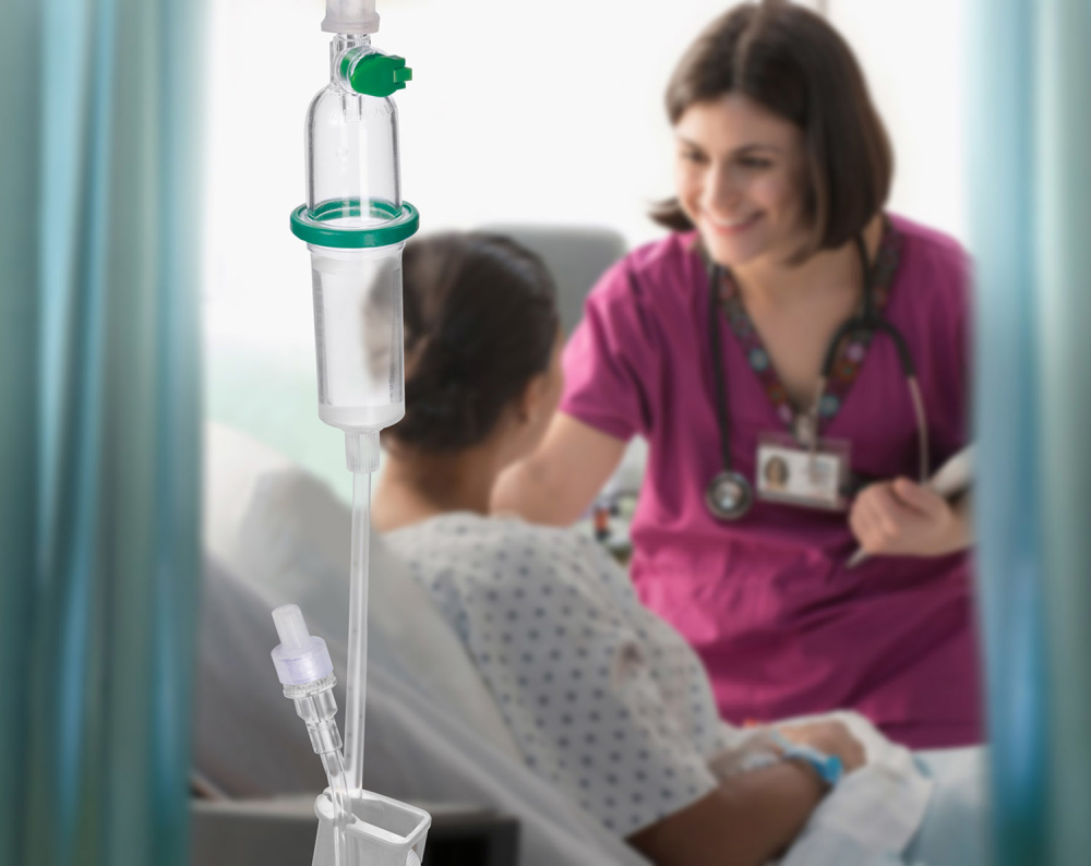 Close-up of the CARESAFE IV Administration Set in the foreground with a blurry healthcare provider smiling and leaning down to speak with a patient in the background.