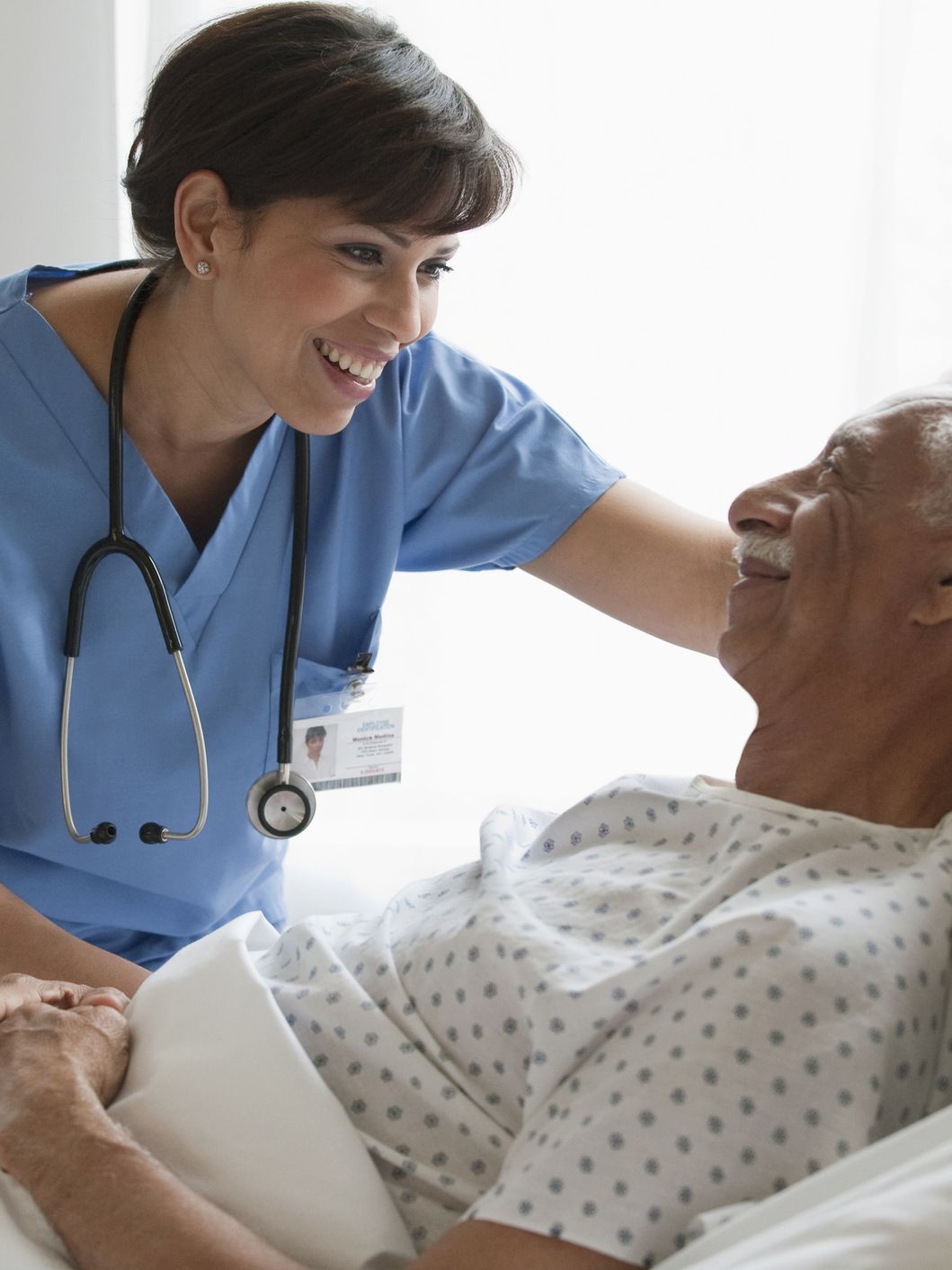 Smiling nurse in scrubs leaning in to greet a male patient in a hospital bed.
