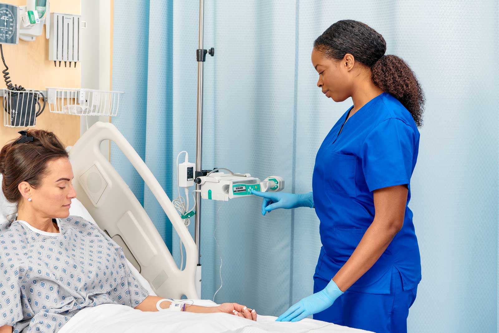 Female patient resting in hospital bed while a nurse programs a Perfusor Space Syringe pump on an IV pole next to the bed.
