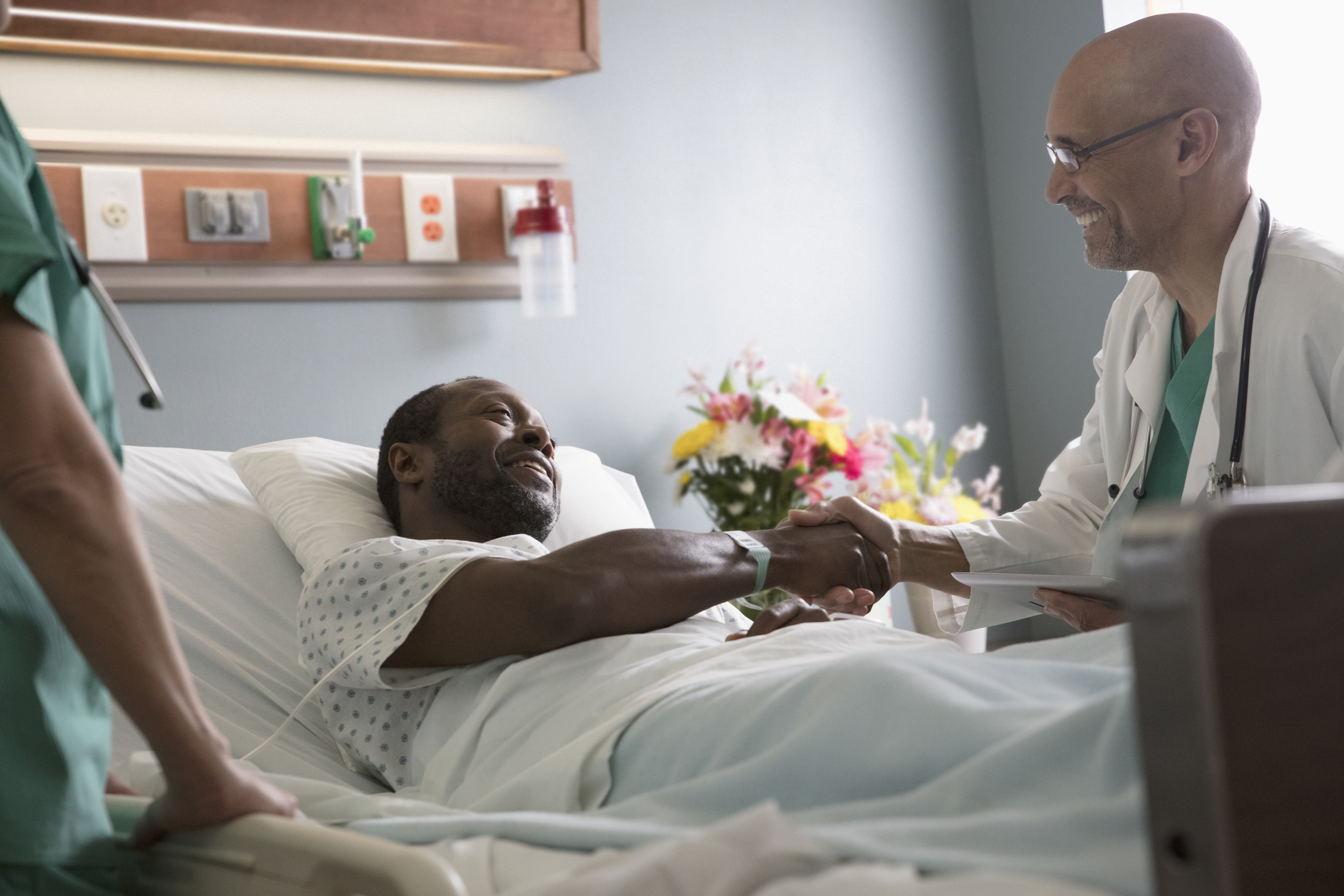 Patient lying in hospital bed smiling and shaking doctors hand who is sitting aside the bed. 