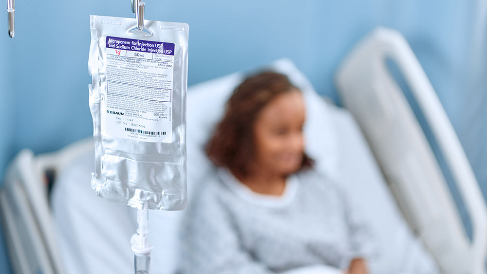 Close-up photo of a B. Braun IV container with a patient sitting in a hospital bed, out of focus, in the background.