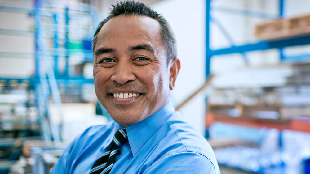Man in a shirt and tie smiling at the camera with warehouse shelving in the background.