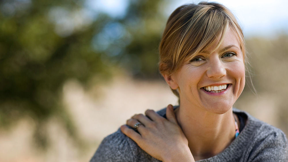 Woman smiling at the camera with one hand on her shoulder while outside.